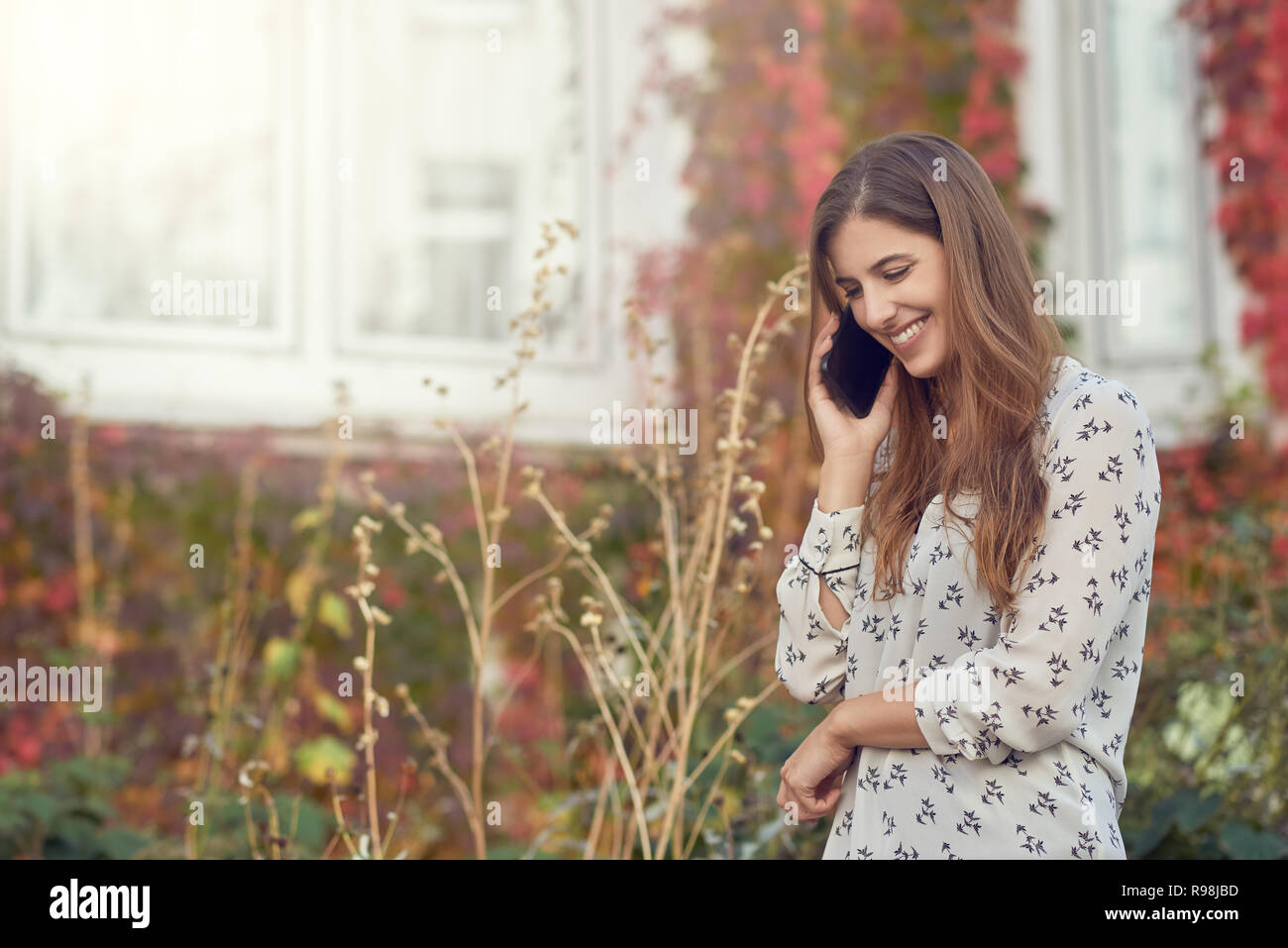 Souriante jeune femme chat sur un mobile à l'automne debout devant une maison blanche avec réducteur colorés smiling Banque D'Images