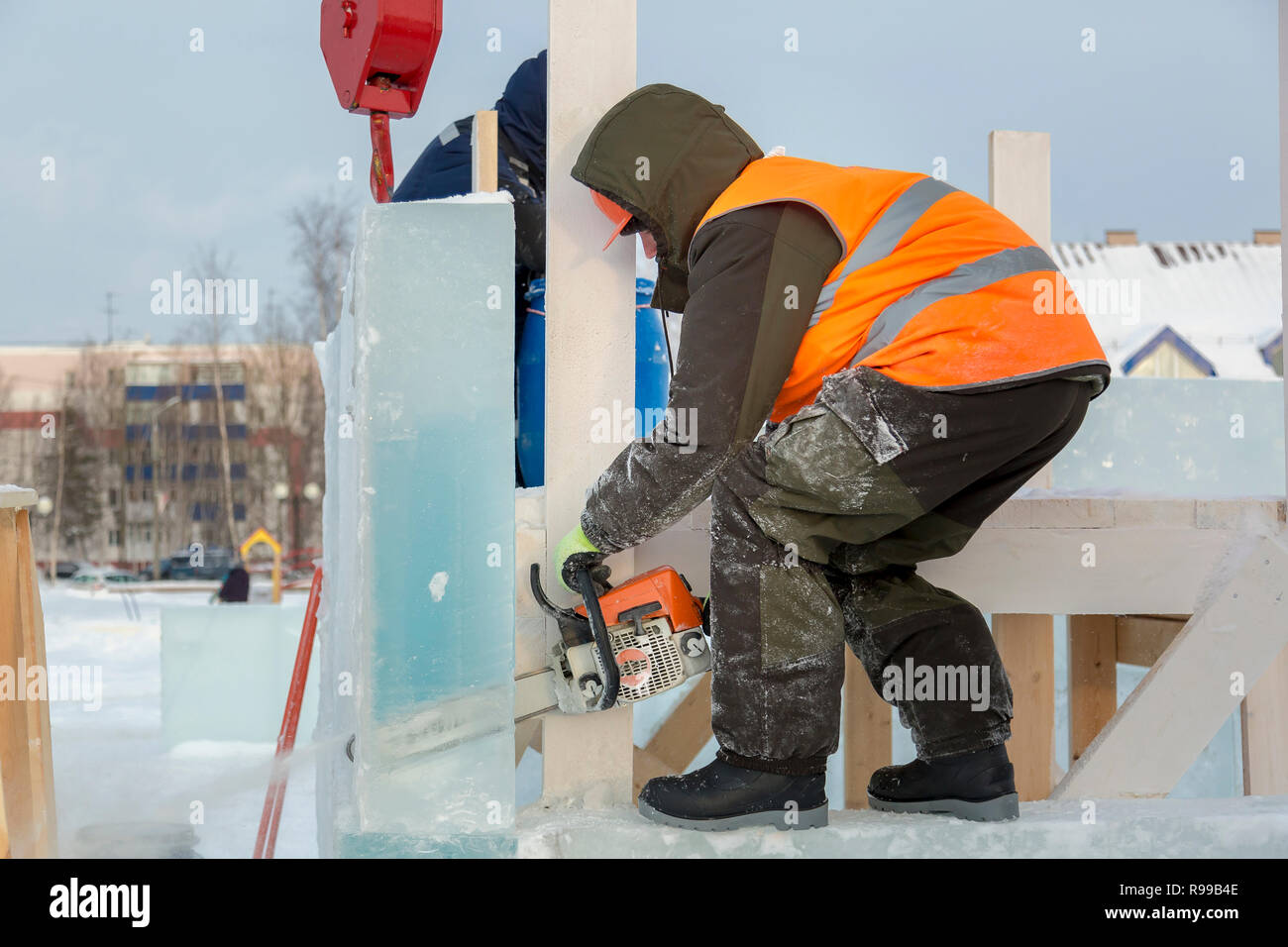 Monteur travailleur dans un casque de protection et gilet réfléchissant une plaque de glace à scier avec une scie à chaîne Banque D'Images