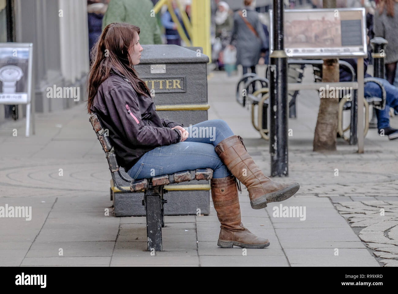 Une jeune femme aux cheveux longs en blouson de cuir, bottes et jeans attend sur un banc dans une zone piétonne du centre-ville urbain shopping area Banque D'Images