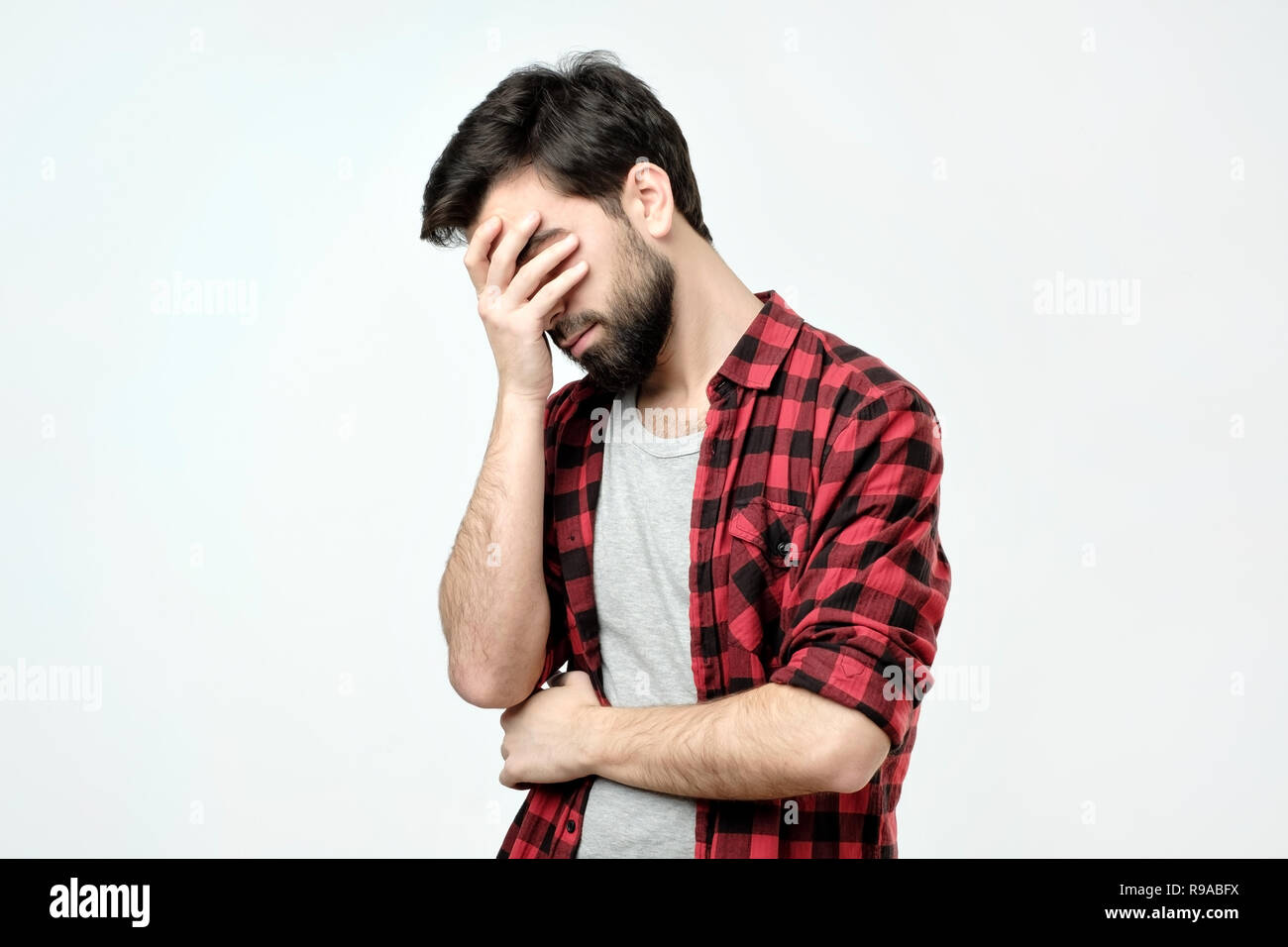 Jeune homme xhausted couvrant le visage avec la main en position debout contre l'arrière-plan gris Banque D'Images