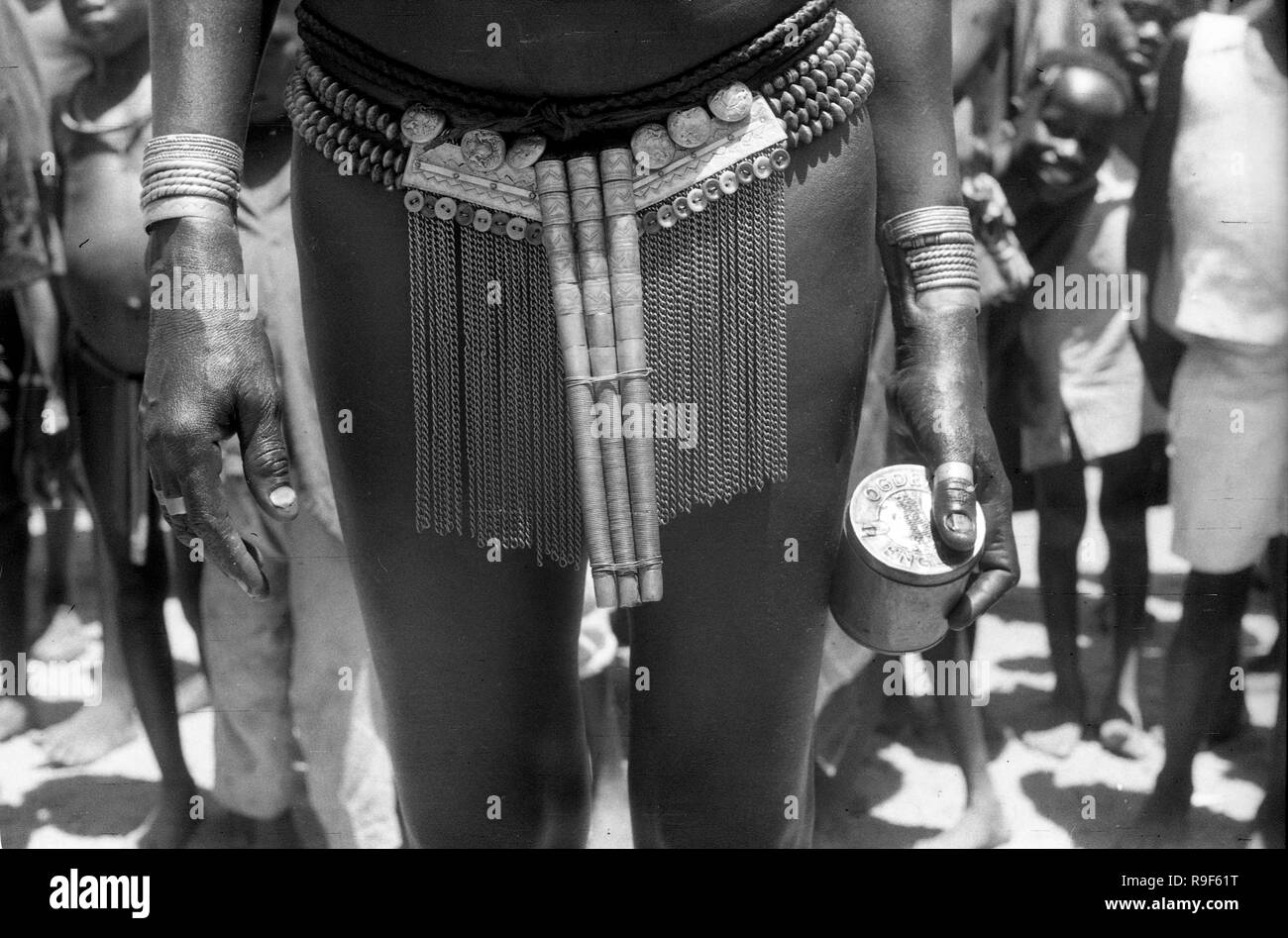 Femme autochtone des années 1950 portant des décorations traditionnelles du corps tribal tout en faisant le commerce et le transport de la boîte à tabac Ogdens au marché de rue en Afrique de l'Ouest Banque D'Images