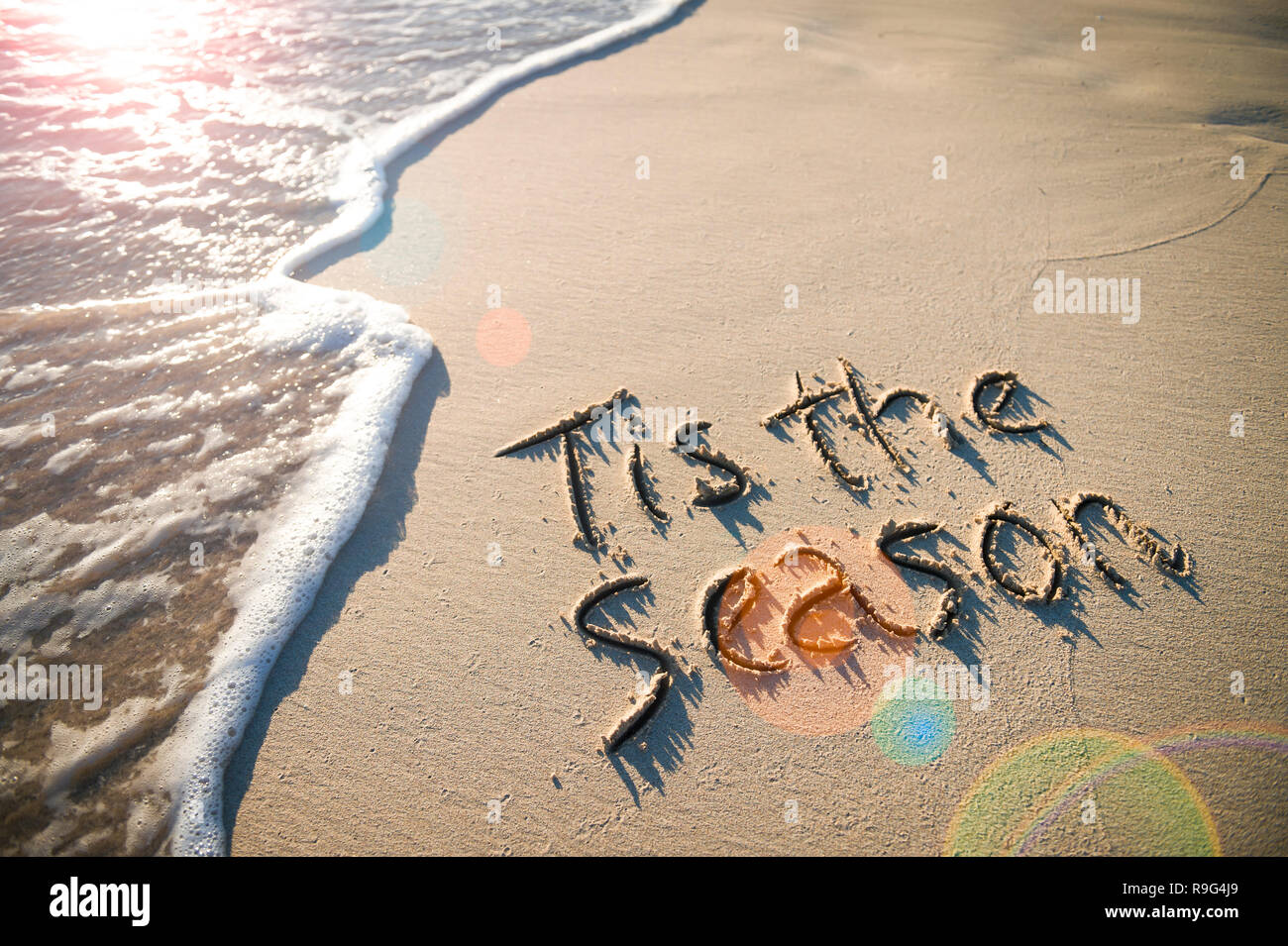 Tis la saison message de vacances de Noël à la main sur une plage de sable fin avec l'arrivée d'un vague sous un soleil matin lens flare Banque D'Images