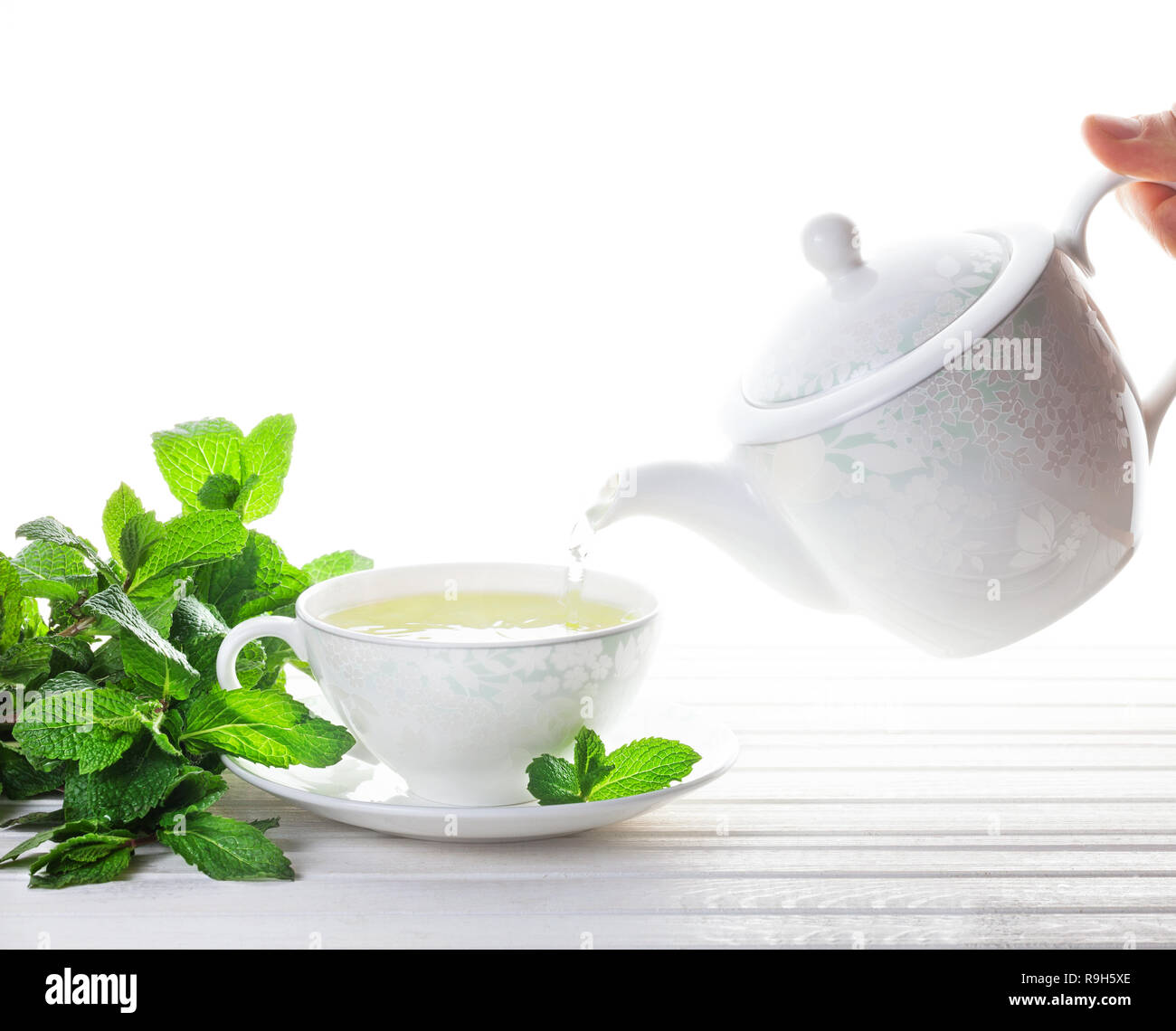 Le thé à la menthe coule à partir de la théière dans la tasse près de feuilles de menthe au fond blanc Banque D'Images