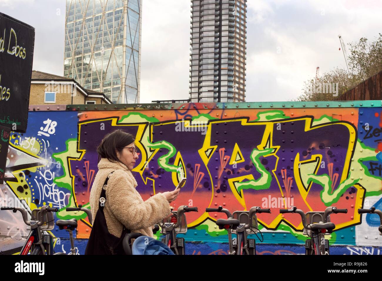 Une femme à la recherche de son téléphone intelligent avec un paysage urbain derrière elle de graffitis et de tours, sur Brick Lane, East London Banque D'Images