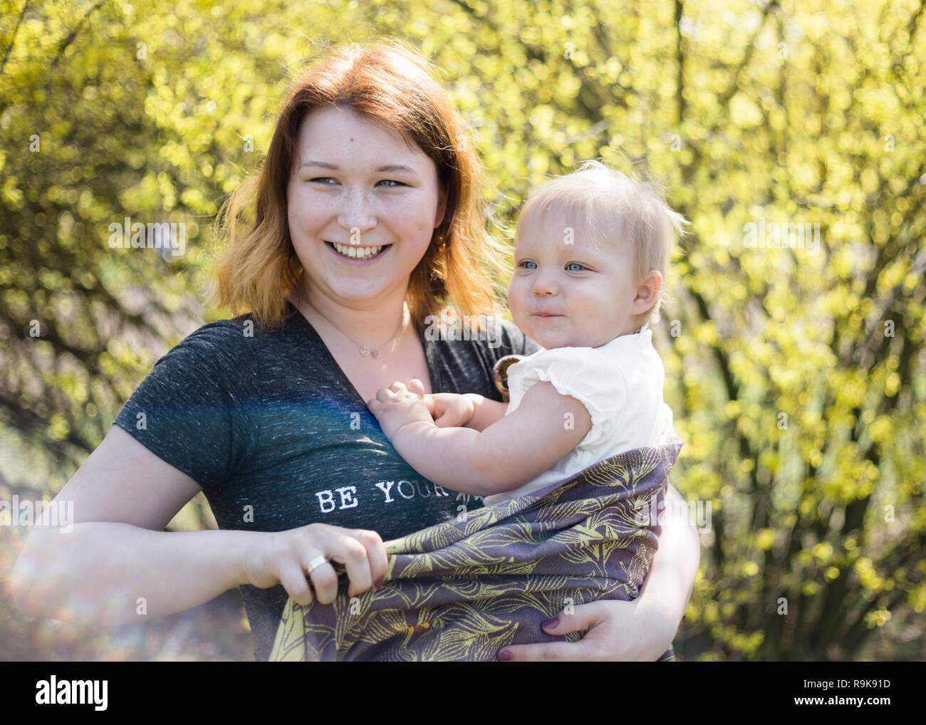 Mother smiling pendant le liage sa petite fille dans une écharpe de portage. Image Style de vie tourné sur place dans un parc sur une journée de printemps ensoleillée. Banque D'Images