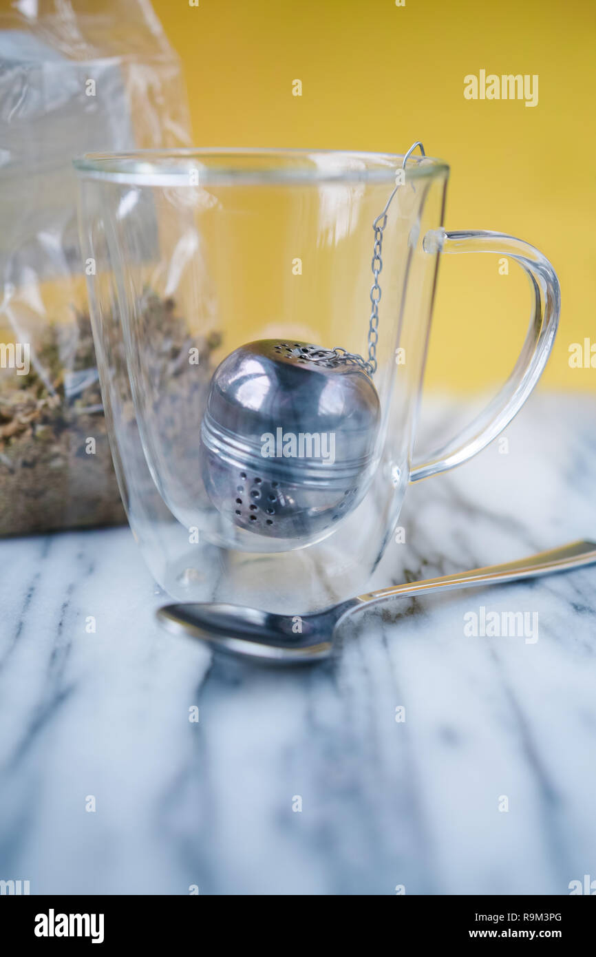 L'intérieur de la tasse en verre de thé Nous avons une boule à thé. Nous voyons également un sac transparent avec fleurs de camomille séchées. Fond jaune. Banque D'Images