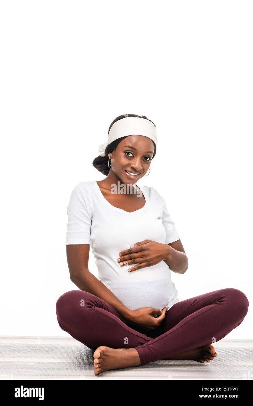 Happy african american pregnant woman sitting on floor et en gardant les mains sur le ventre isolated on white Banque D'Images