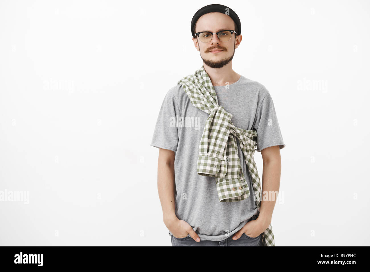 Studio shot de calme et froid creative européenne homme avec barbe, dans les verres et black beanie holding hands in pockets ricanant et contemplant avec indifférence à huis clos autour de l'ennui Banque D'Images