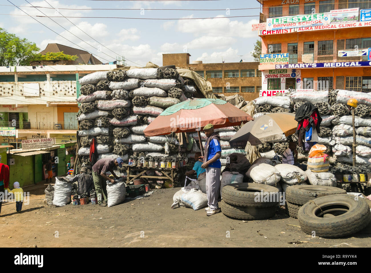 Grand charbon sacs empilés à côté d'une route par des bâtiments avec des hommes sacs de remplissage à l'avant, au Kenya Banque D'Images