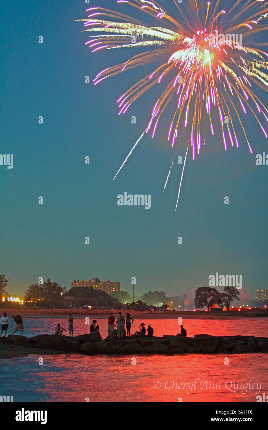 Groupe de personnes artifice à partir d'une jetée. La réflexion d'artifice s'allume en rouge dans l'eau autour d'eux. Banque D'Images