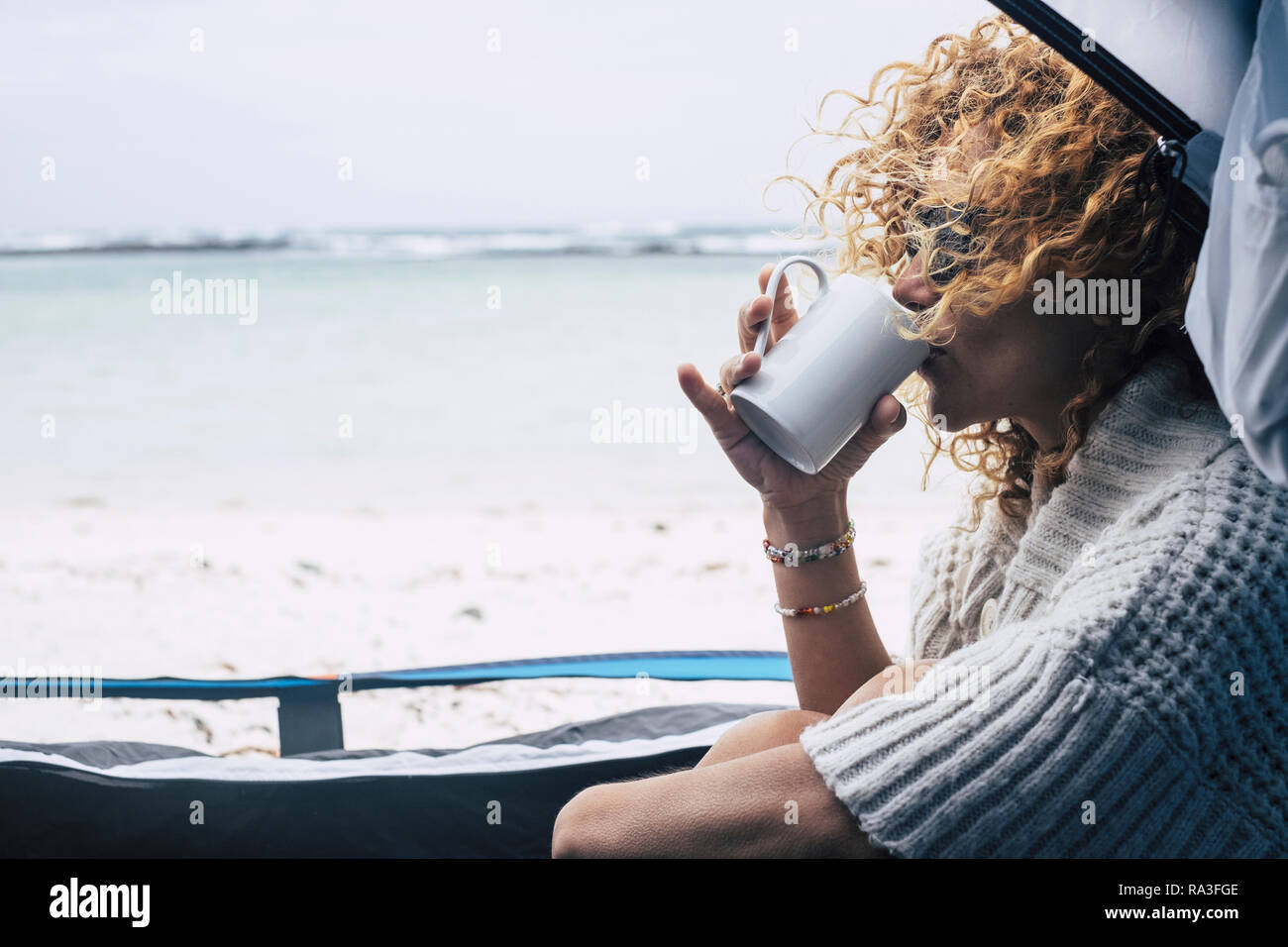 Belle femme d'âge moyen s'asseoir à l'intérieur d'une tente en plein air camping plage libre de sentir la nature tandis que boire un thé ou un café dans une tasse blanche - vent Banque D'Images