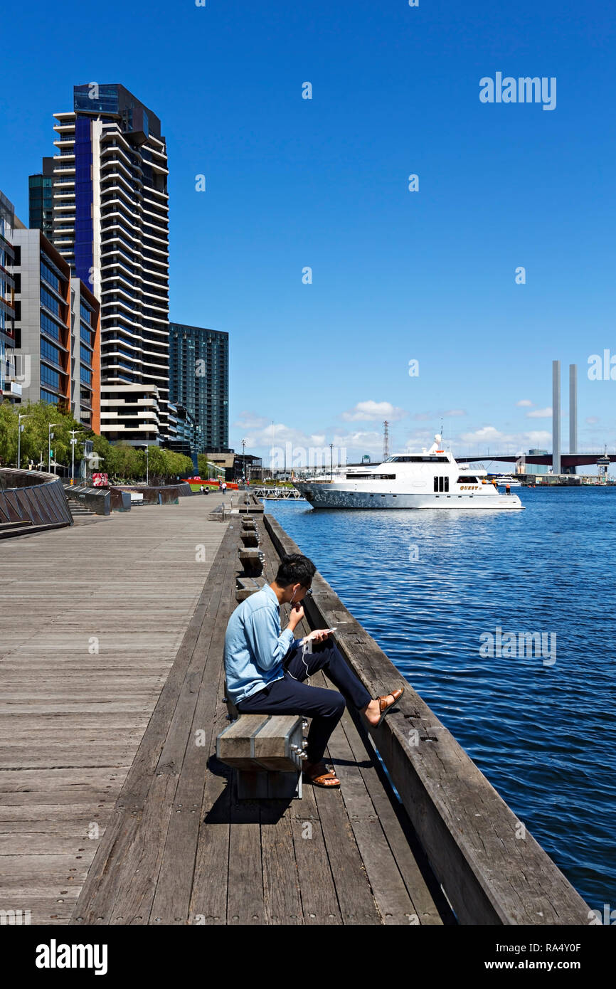 Un jeune homme asiatique est à l'écoute de la musique sur son téléphone portable dans le port de Victoria à Melbourne Docklands,Victoria en Australie. Banque D'Images