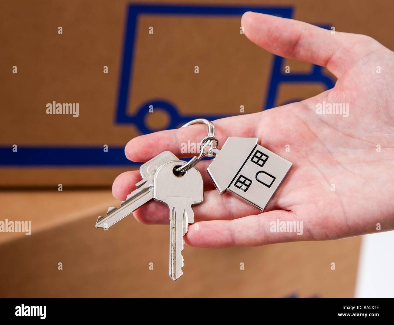 Main tenant des clés de la maison avec une clé en métal en forme de tag comme une maison, à l'arrière des boîtes de déménagement Banque D'Images