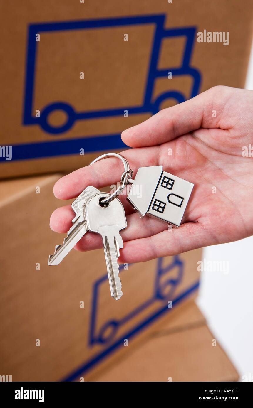 Main tenant des clés de la maison avec une clé en métal en forme de tag comme une maison, à l'arrière des boîtes de déménagement Banque D'Images