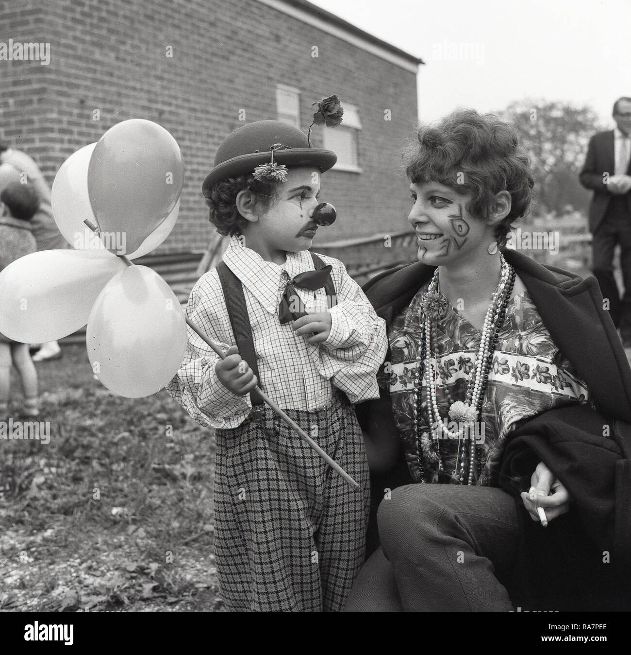 1965, fête du village, la mère avec le mot 'Amour' peint sur son visage, avec elle, un jeune garçon en robe de soirée, portant un chapeau et le nez d'un clown et ballons holding, England, UK. Banque D'Images