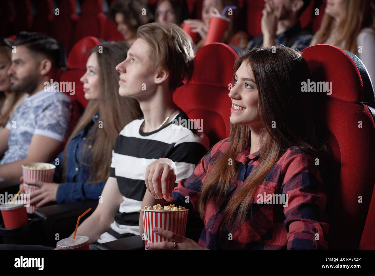 Voir du côté de la belle jeune fille aux cheveux noirs en chemise rouge profiter regarder des films intéressants au cinéma. Jolie femme spectateur assis près de garçon blond et eating popcorn de red cup. Banque D'Images