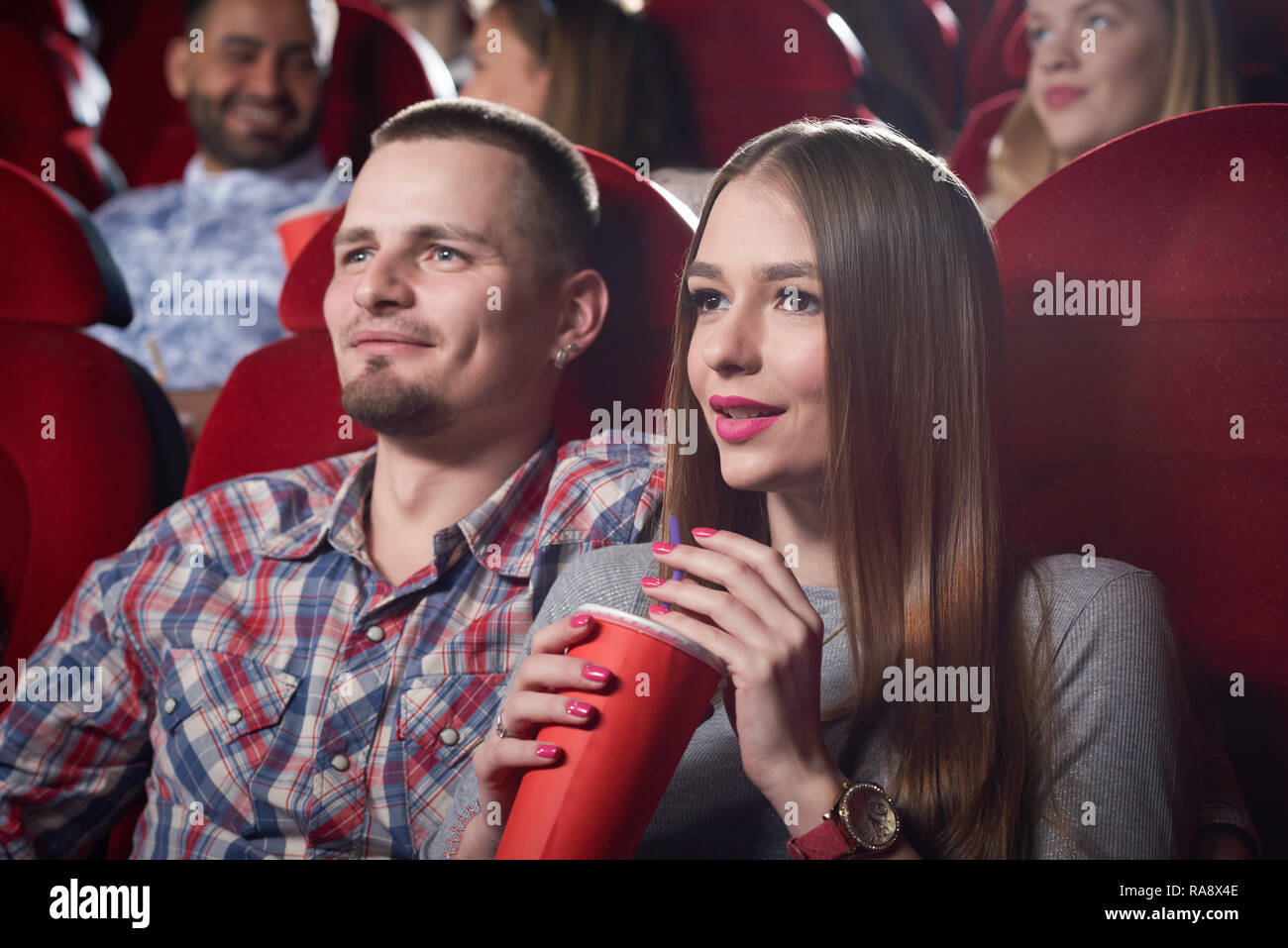 Couple de l'homme en chemise à carreaux et la femme en gris assis ensemble dans le cinéma, regarder film romantique. Beau barbu hugging belle petite amie qui assis près de holding et tasse de boisson. Banque D'Images