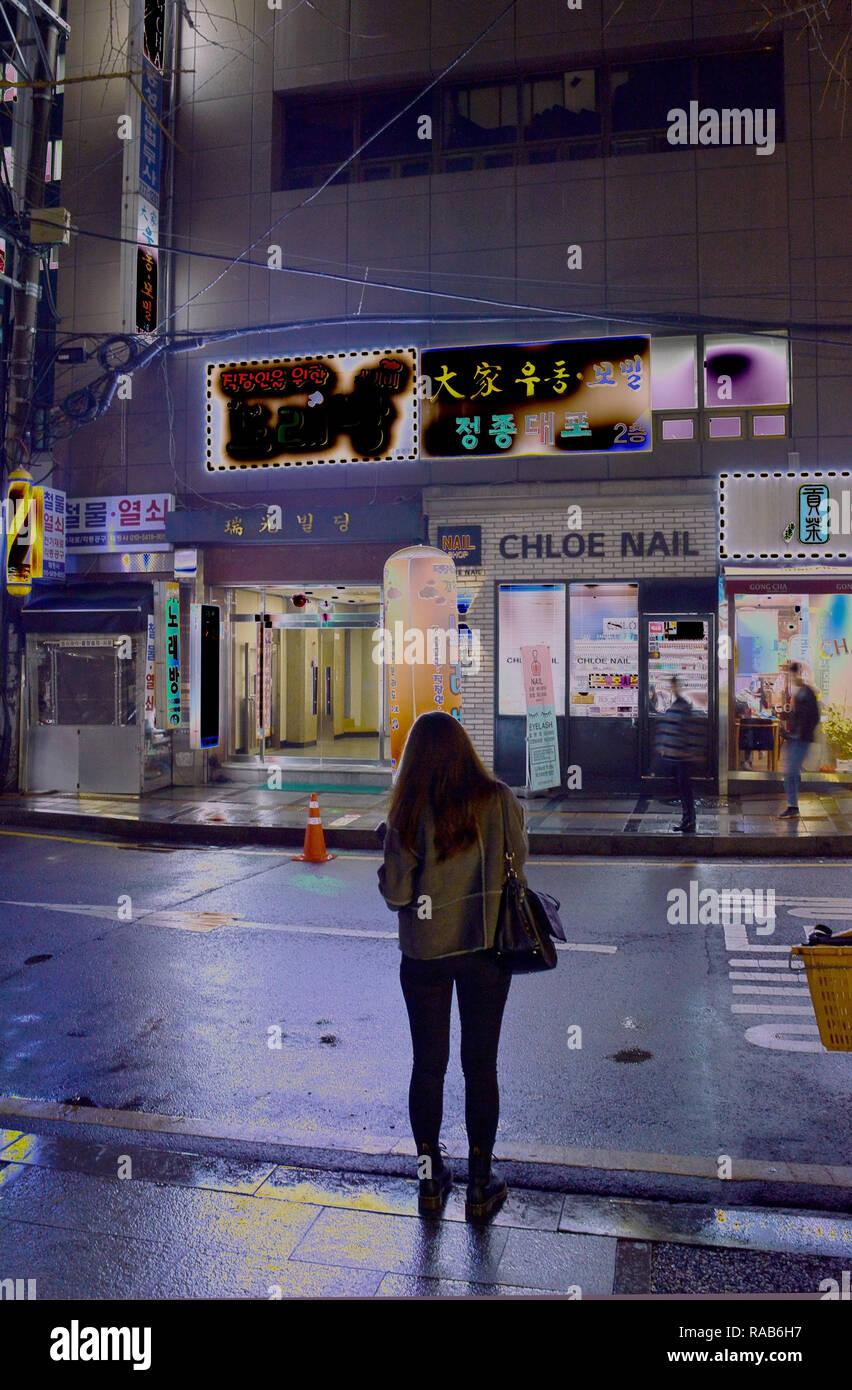 Un long-haired woman stands, dos à la caméra, se découpant sur le bord d'une rue de ville baignée de lumière néon à Séoul, Corée du Sud Banque D'Images