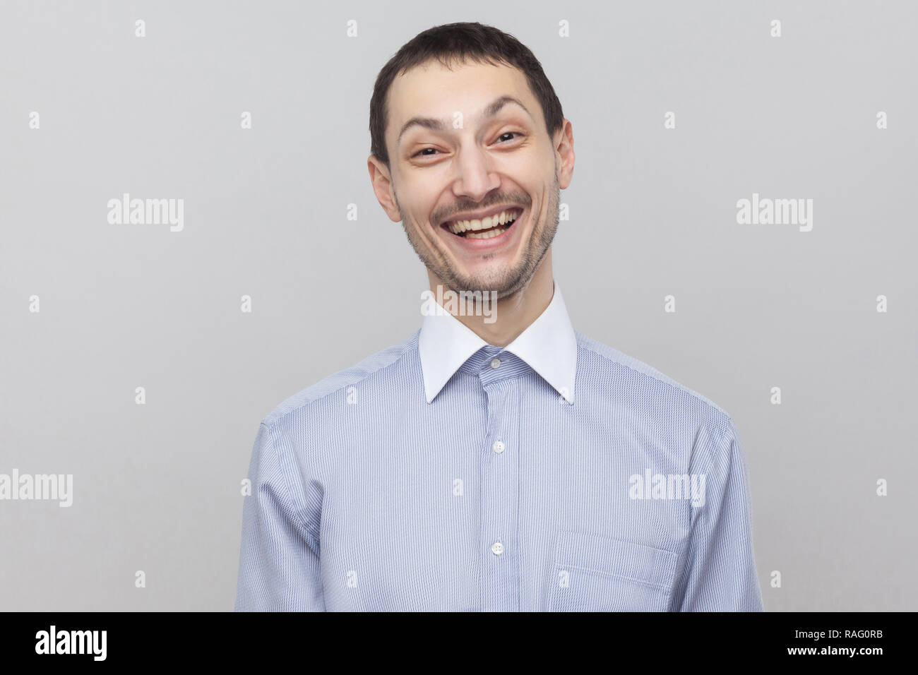 Portrait of happy young en soies beau satisfait classic shirt bleu clair et permanent à la recherche à l'appareil photo avec drôle de visage, rire intérieur. Banque D'Images Portrait of happy young en soies beau satisfait classic shirt bleu clair et permanent à la recherche à l'appareil photo avec drôle de visage, rire intérieur. Banque D'Images