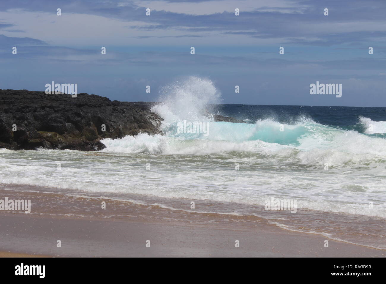 Éclaboussures des vagues de l'océan près de la côte de black rock au cours de l'été à une plage à Kauai, Hawaï. Il y a quelques nuages gris dans le ciel. Banque D'Images