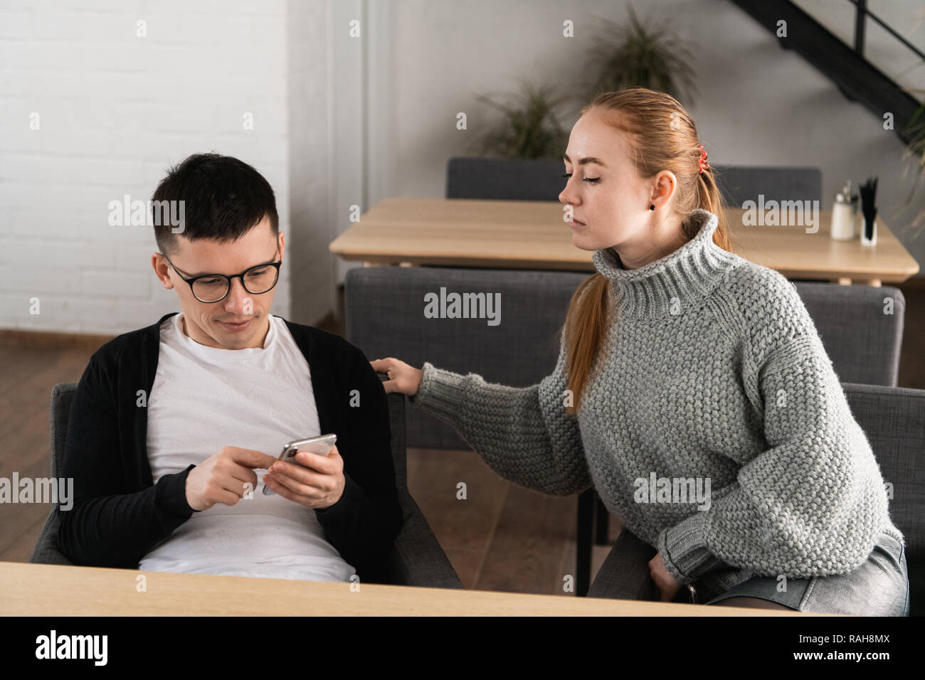 Cheater homme recherchant à la ligne avec un téléphone intelligent et petite amie d'espionnage est assise sur un canapé dans le café Banque D'Images