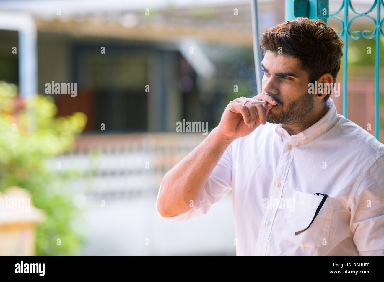 Beau jeune homme indien barbu pense en fumant à l'extérieur Banque D'Images