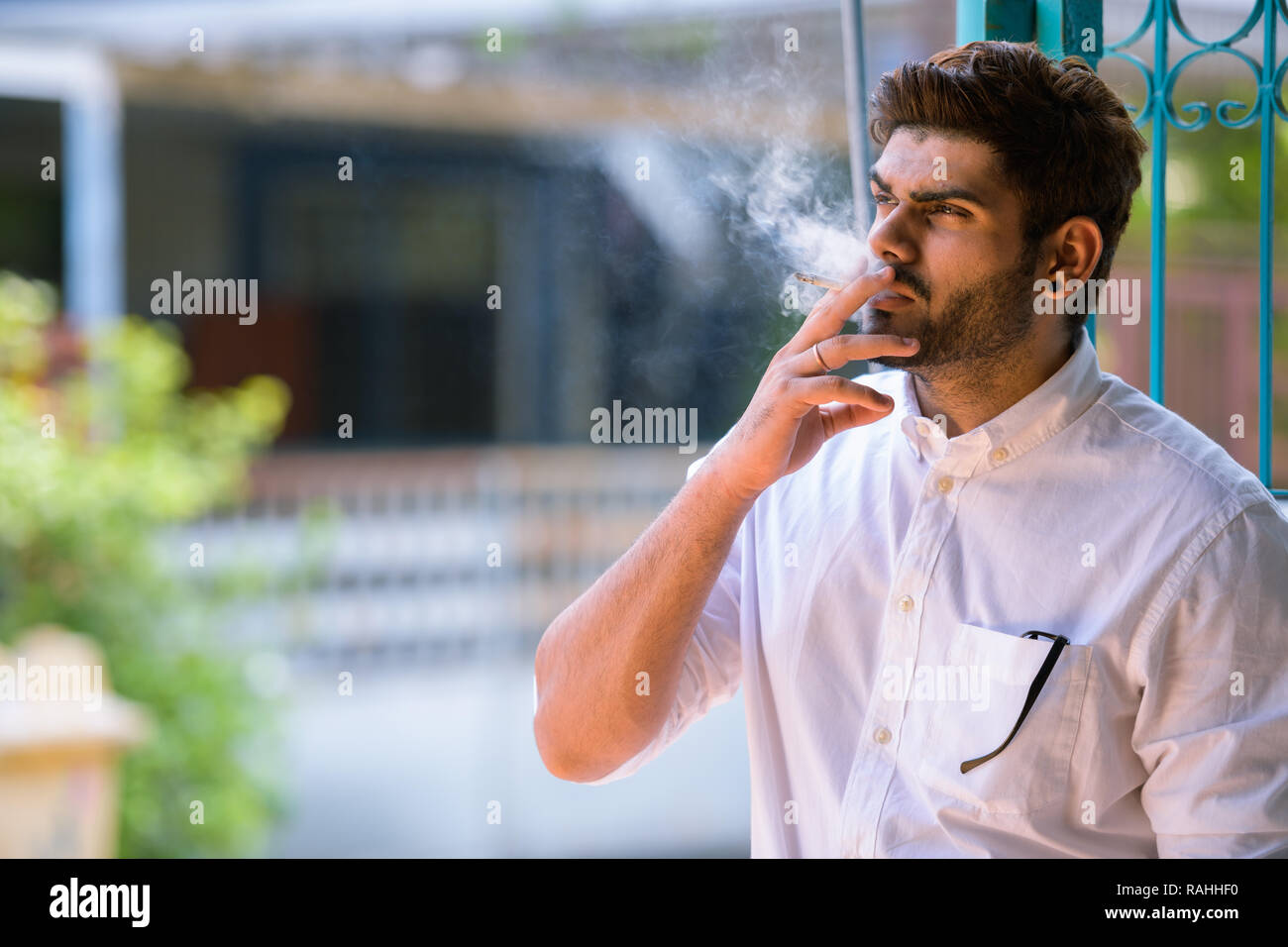 Beau jeune homme indien barbu pense en fumant à l'extérieur Banque D'Images
