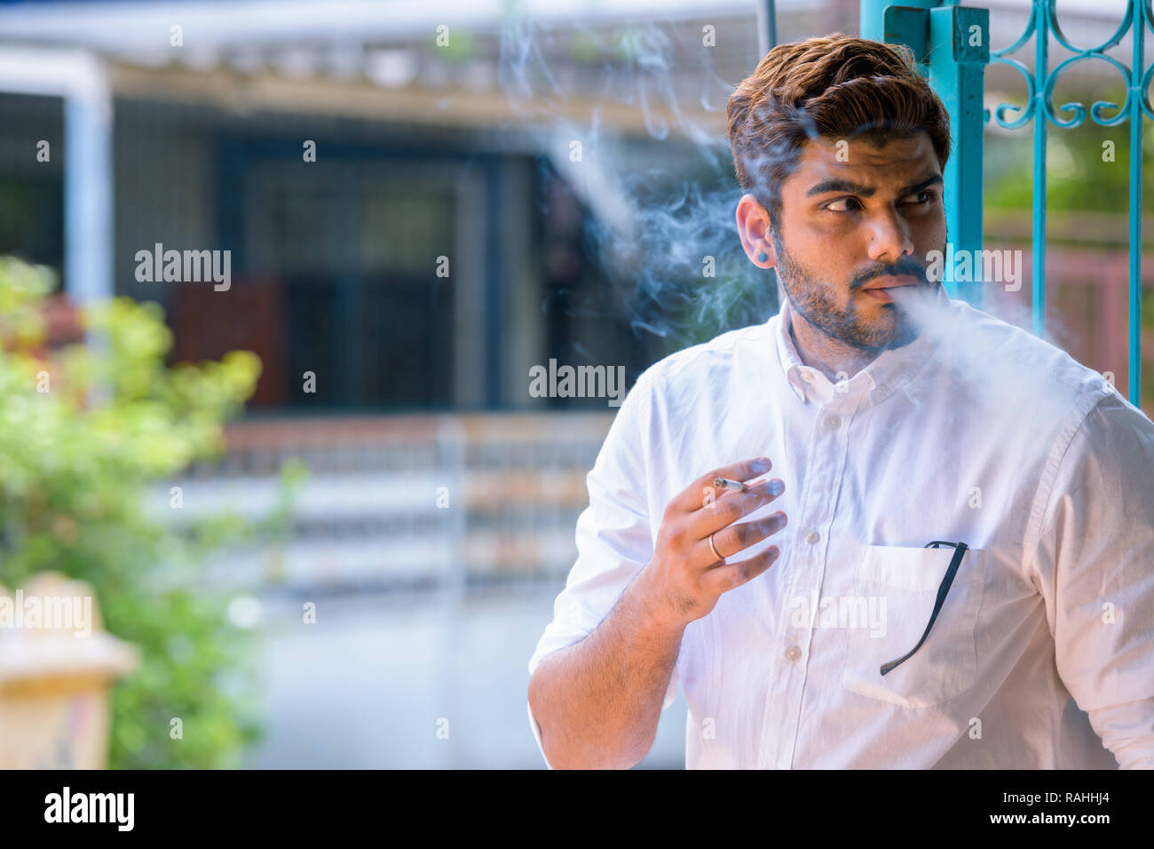 Jeune homme indien barbu pense en fumant cigarette à l'extérieur Banque D'Images