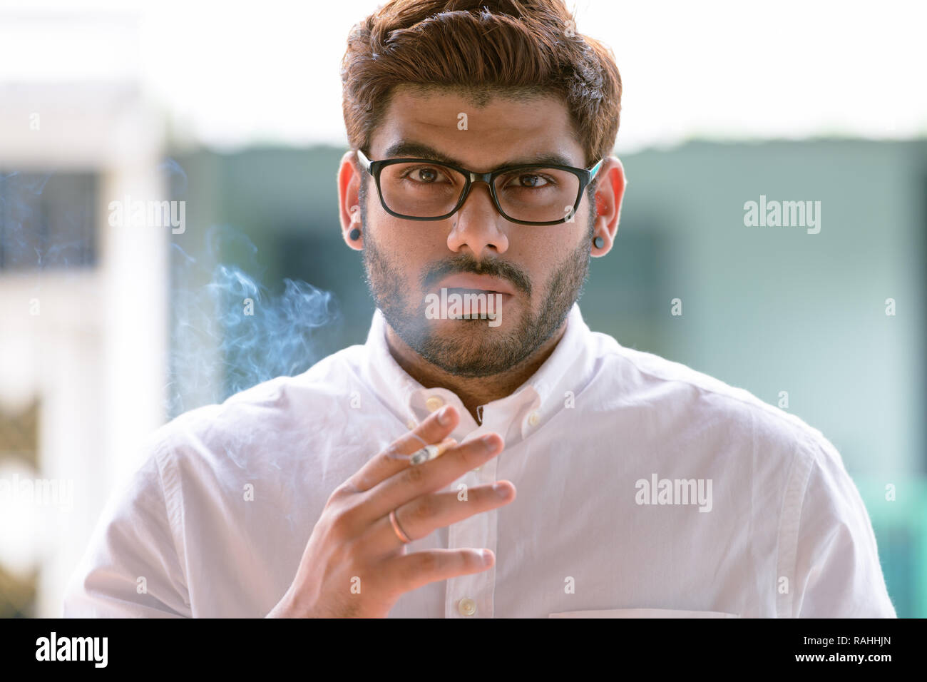Visage de jeune homme barbu avec des lunettes fumeurs à l'extérieur Banque D'Images