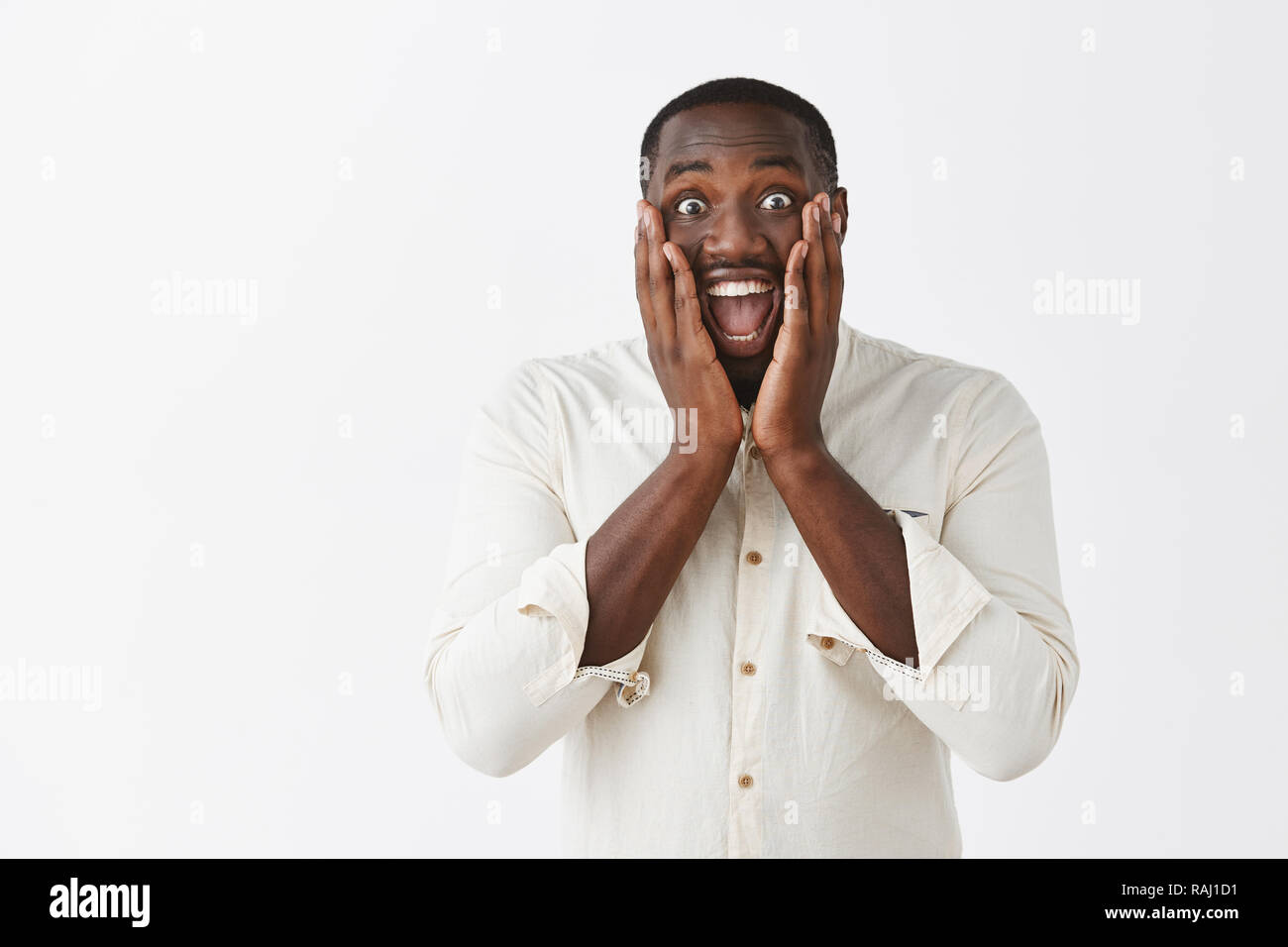 L'homme à l'audience d'une excellente nouvelle, d'être joyeux et émerveillés, heureux pour ami. Portrait d'impressionné et surpris optimiste African American gars en chemise blanche et large sourire holding paumes sur les joues Banque D'Images