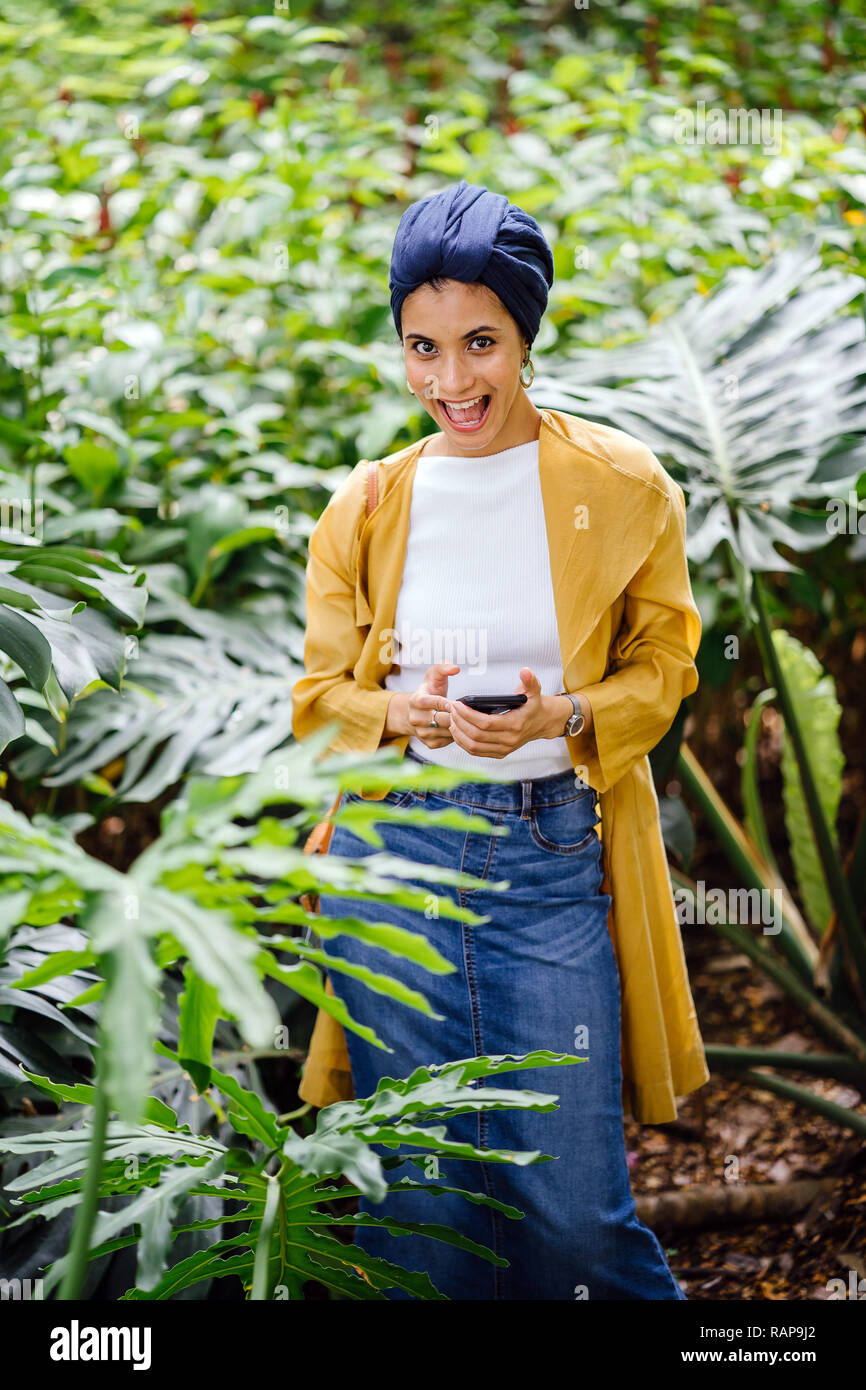 Portrait d'une jeune et jolie asiatique malais femme musulmane dans un turban hijab foulard debout dans un parc et sourire comme elle utilise son smartphone. Banque D'Images