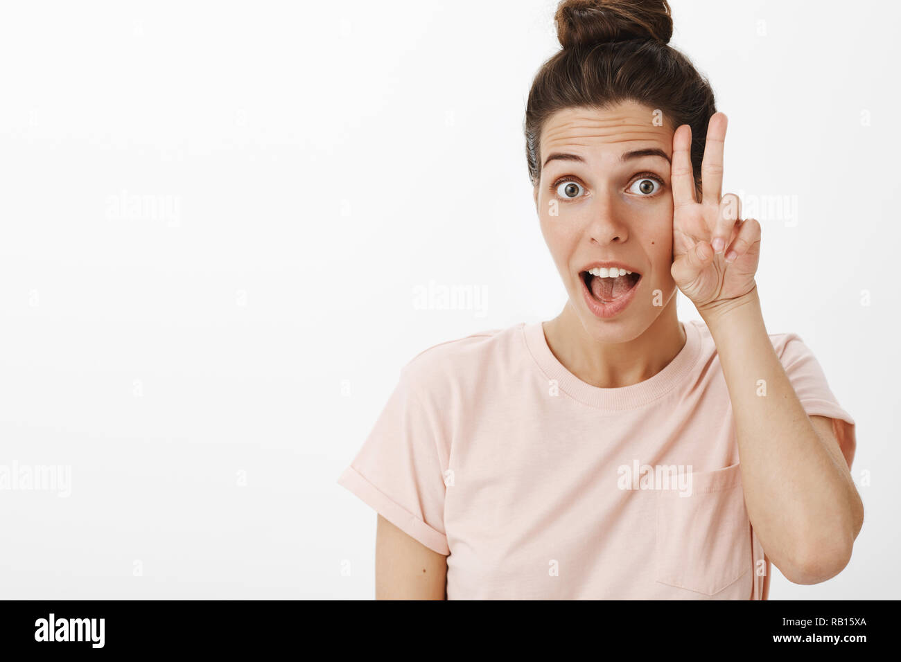 Hey chill et profiter de la vie. Bien insouciante et détendue de l'élégant charismatique woman in pink t-shirt sensibiliser de façon ludique les sourcils montrant comme signe de la paix près de joue, bouche ouverte dans son oui Banque D'Images