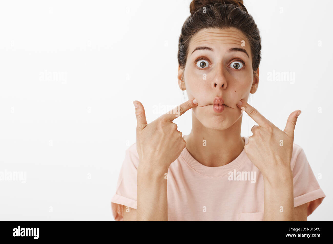 Taille plan sur surpris cute woman étant amusé comme sucer les lèvres et la bouche du poisson en poussant des joues et les sourcils yeux popping dans l'étonnement, de jouer contre l'arrière-plan gris Banque D'Images