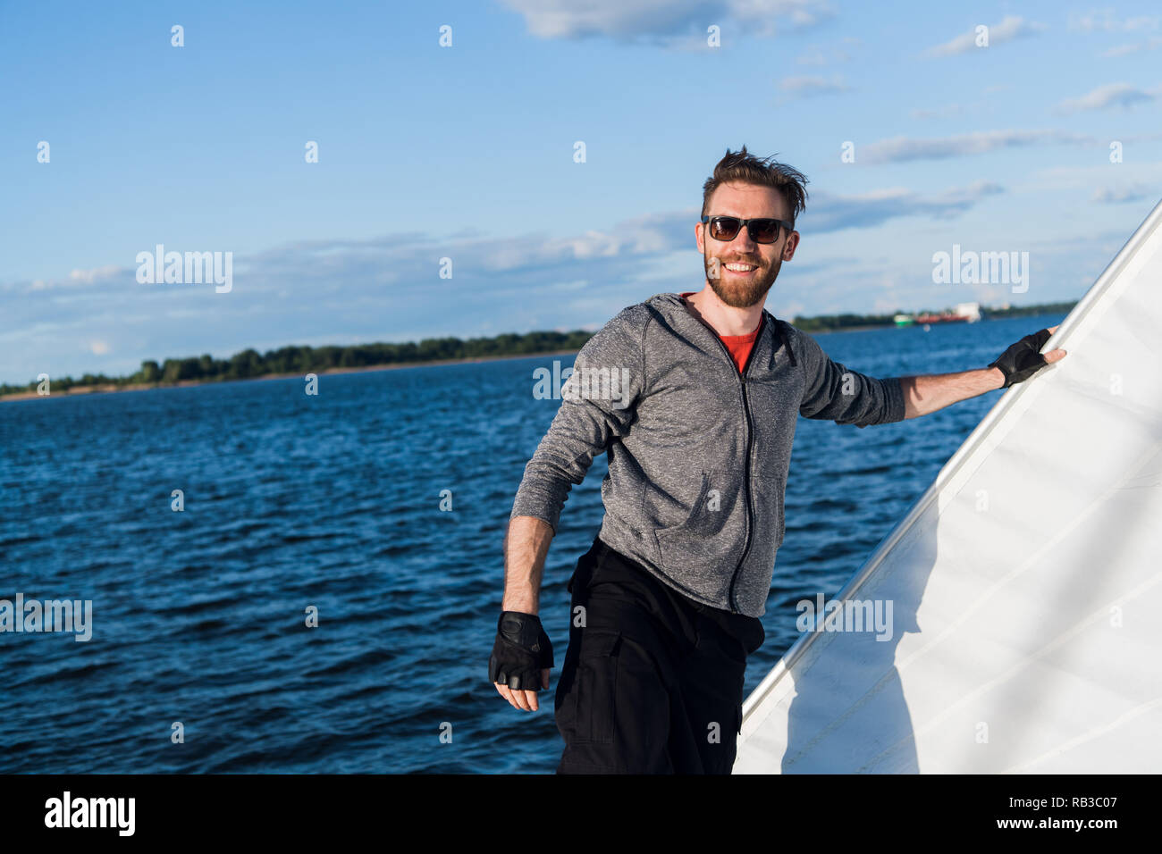 Bel homme voyageant par bateau. Tenue décontractée, l'homme vêtu de gris à capuche, printemps, barbu à lunettes sur le bateau. Banque D'Images