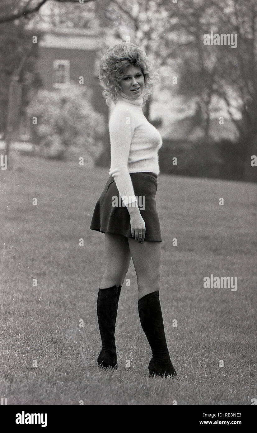 Années 1960, plantureuse femme avec grande les cheveux ondulés et portant une mini-jupe, du cou de tortue et des bottes de cuir posant dehors, England, UK. Banque D'Images