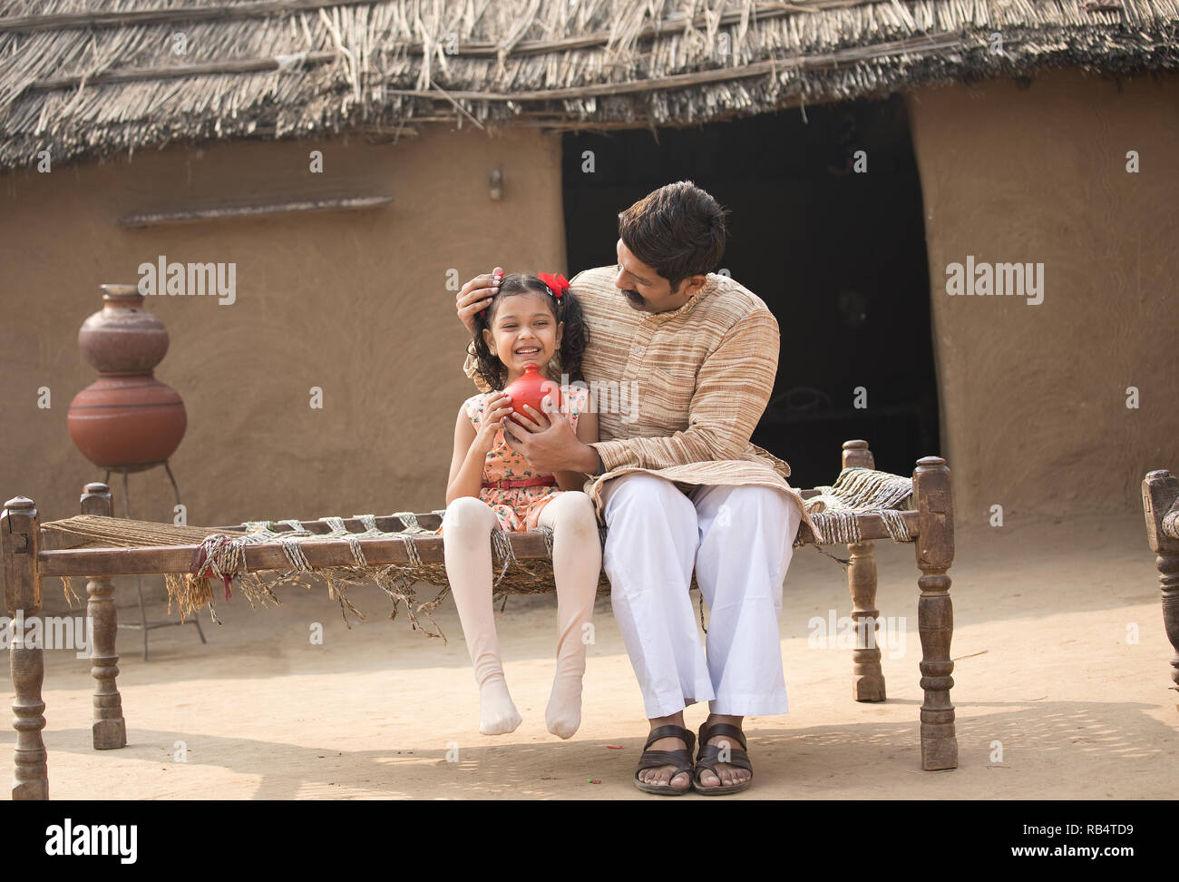 Père indien avec fille holding piggy bank, assis sur un lit traditionnel dans le village Banque D'Images