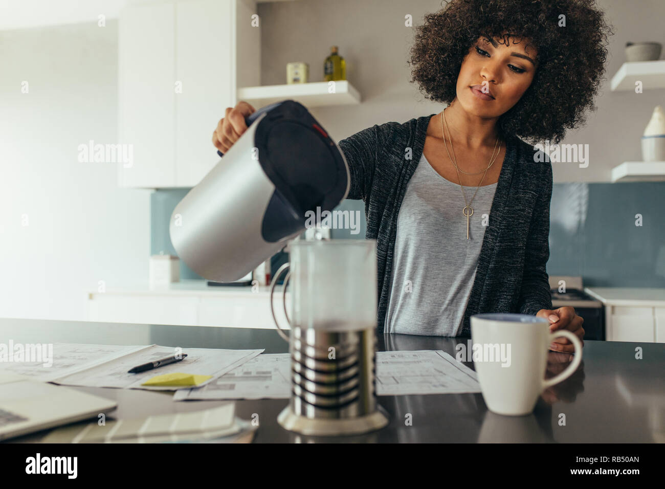 Jeune femme assise à sa table de travail verser de l'eau dans une cruche. Femme africaine à faire le café pour son autonomie tout en travaillant au bureau à domicile. Banque D'Images