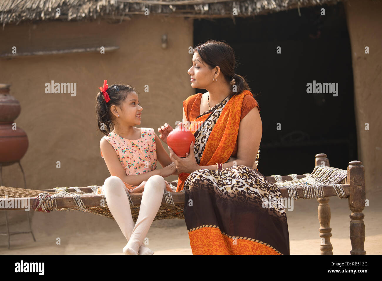 Mère indienne avec fille holding piggy bank, assis sur un lit traditionnel dans le village Banque D'Images