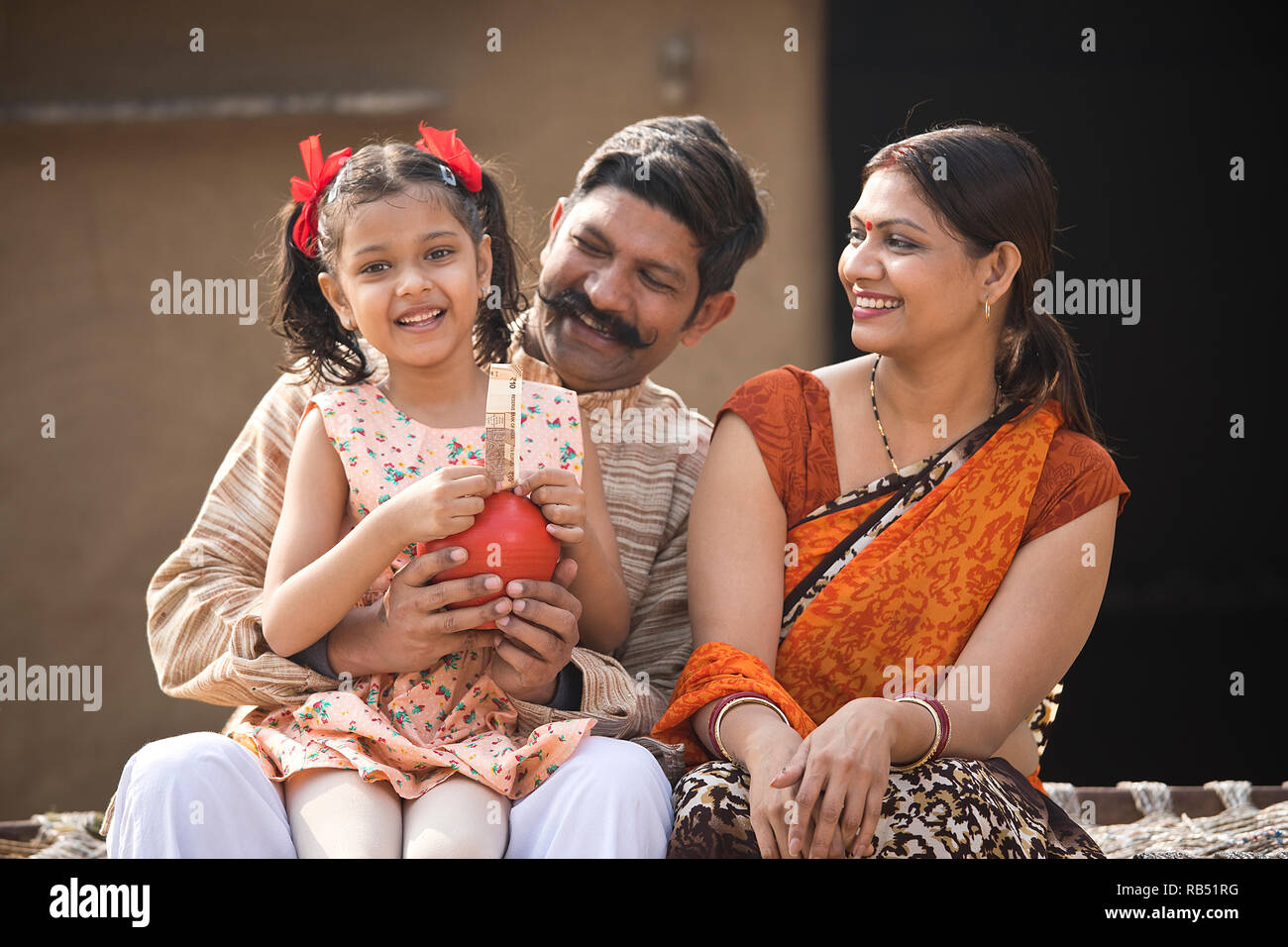 Les parents indiens avec fille holding piggy bank, assis sur un lit traditionnel dans le village Banque D'Images