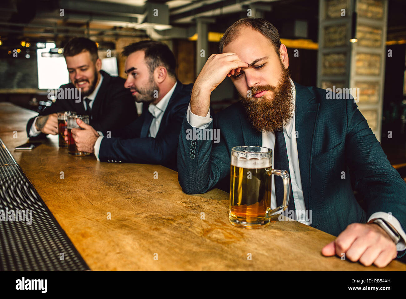 Contrarié et fatigué jeune homme s'asseoir t bar comptoir. Il regarder vers  le bas et maintenez-la main sur le front. Mug stand de bières sur le  comptoir du bar. Deux autres jeunes