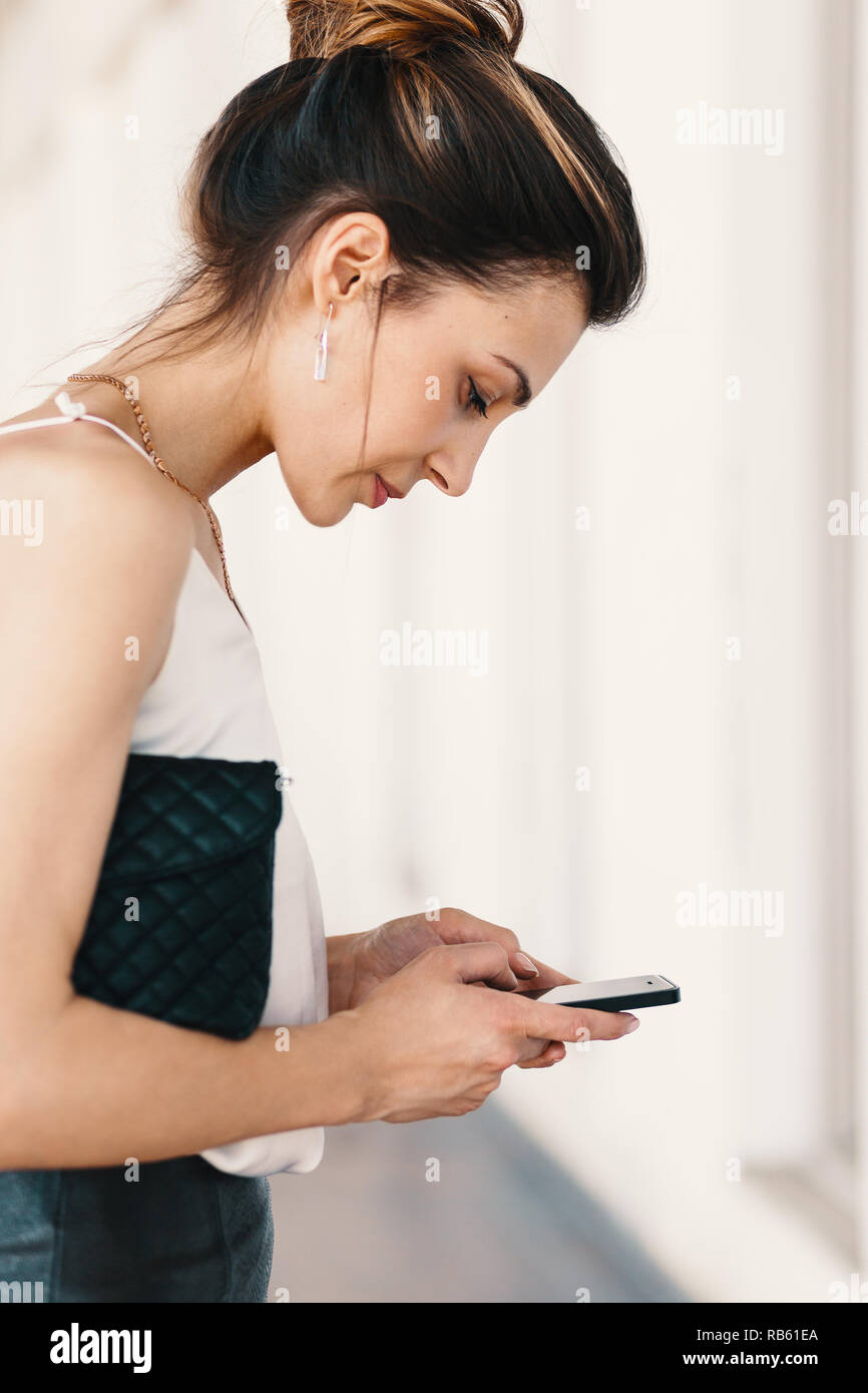 Side view portrait of a smiling, élégante jeune femme utilisant un  téléphone intelligent tout en maintenant un plaid noir à la recherche  d'embrayage au mobile de toucher l'écran avec Photo Stock -