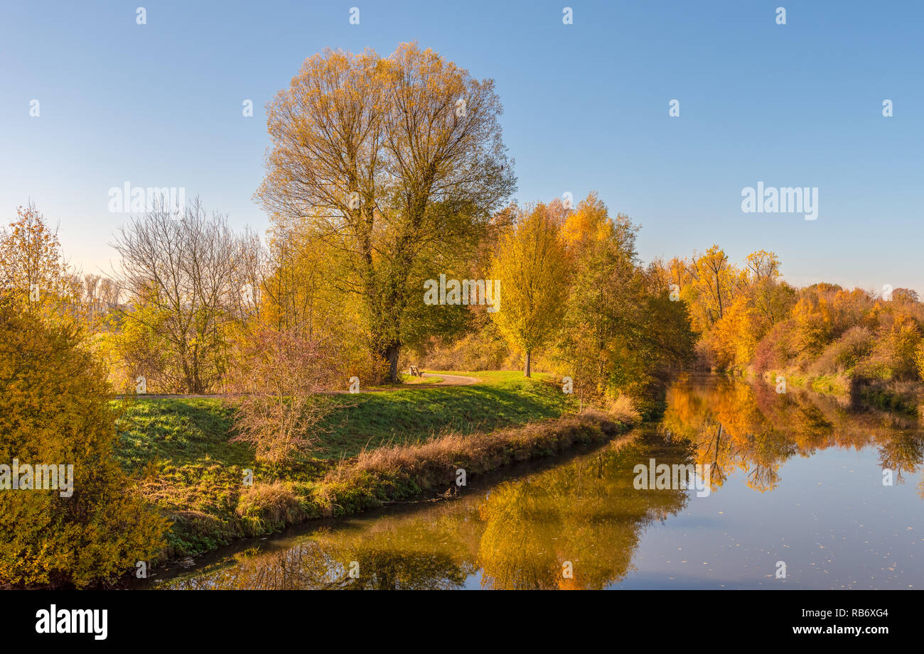 Extérieur couleur automne nature image d'une scène de rivière d'automne rural avec ciel bleu, coloré arbres se reflétant sur l'eau, un banc et un chemin Banque D'Images