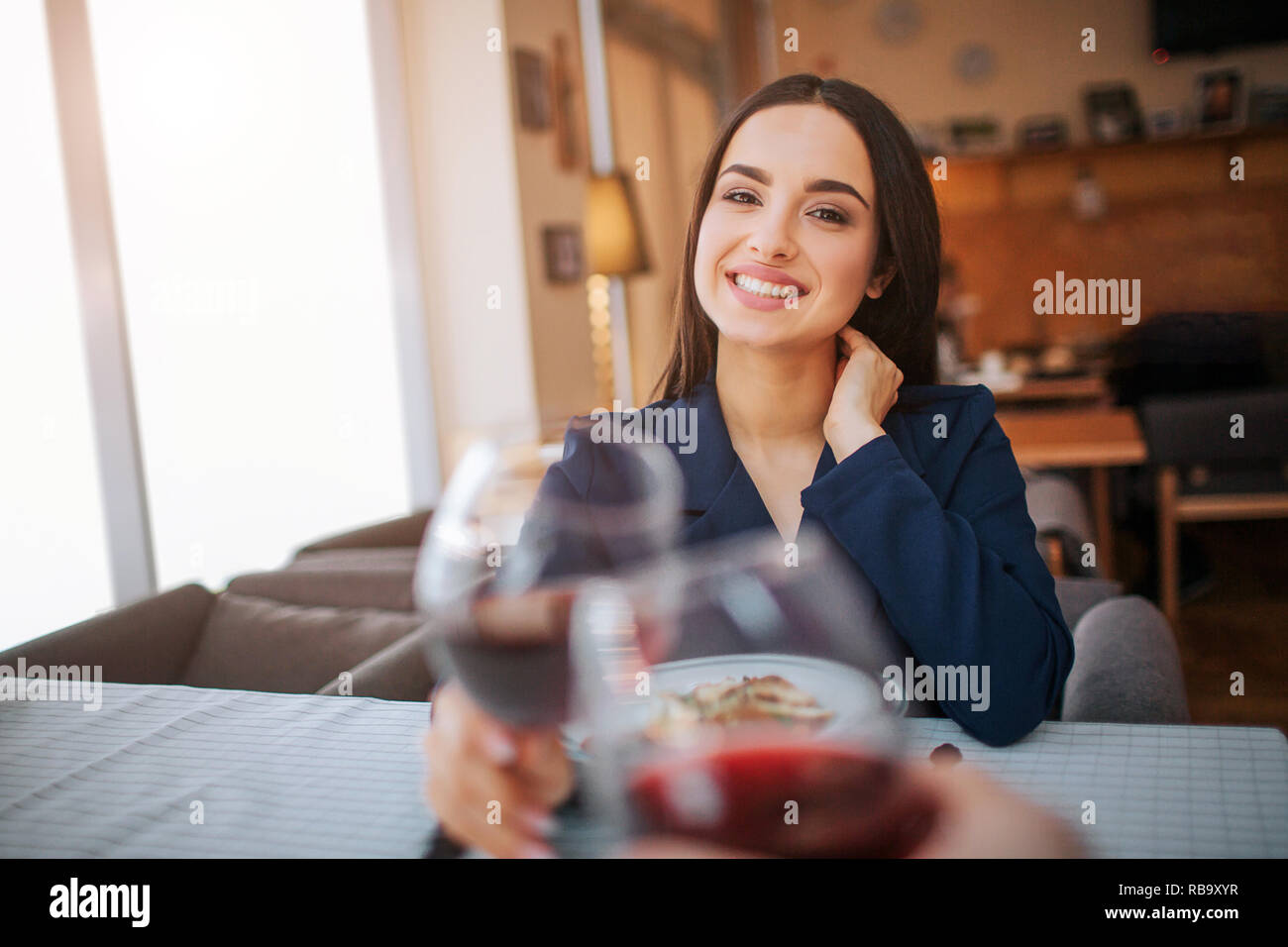 Cheerful yougn femme maintenir le verre de vin rouge et le toucher avec un autre. Elle sourire et regarder droit devant Banque D'Images