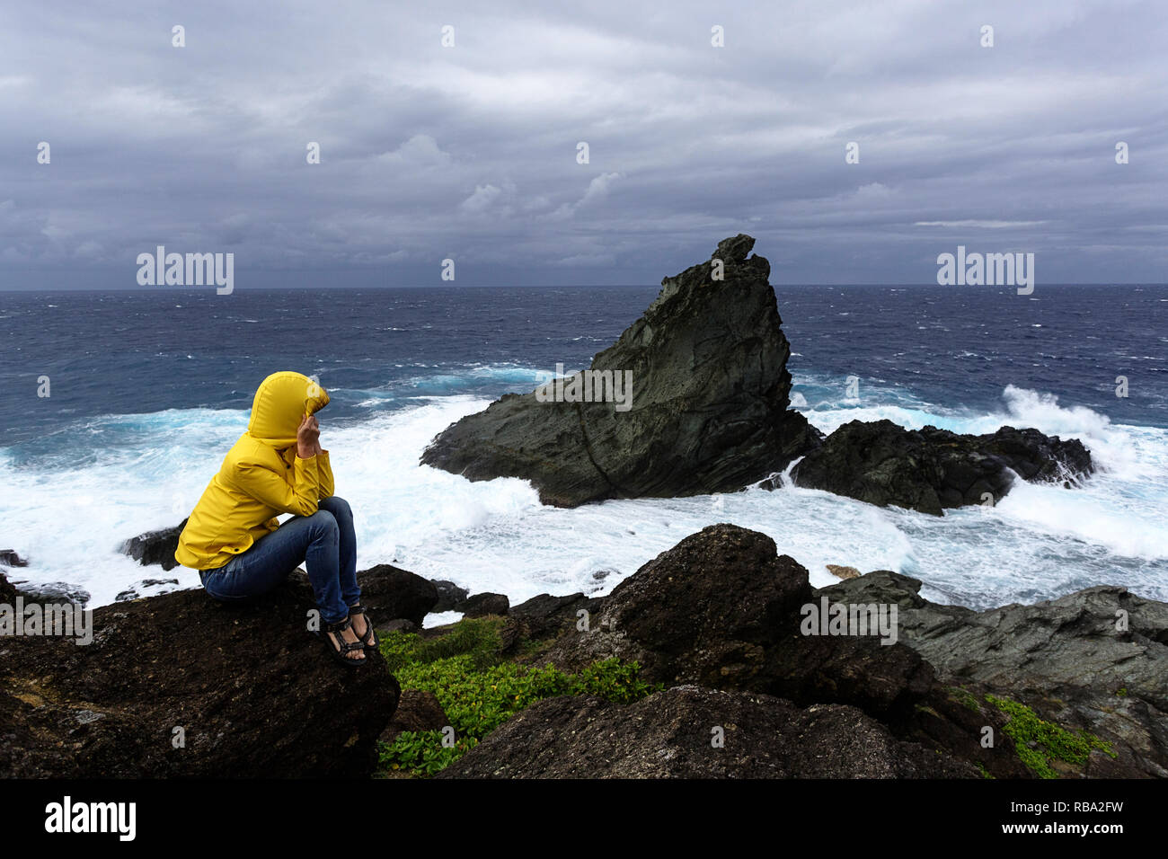 Woman in yellow jacket gel sur un rocher à la recherche à l'état de la mer sur la côte sauvage à Uganzaki dans le phare de l'Île Ishigaki, Japon Okinawa Banque D'Images