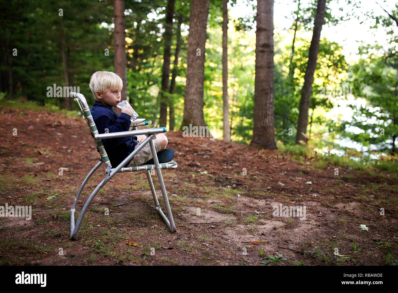 Un mignon petit Blond kid est boire du jus qu'il est assis dans une chaise de jardin vintage en camping près du lac. Banque D'Images