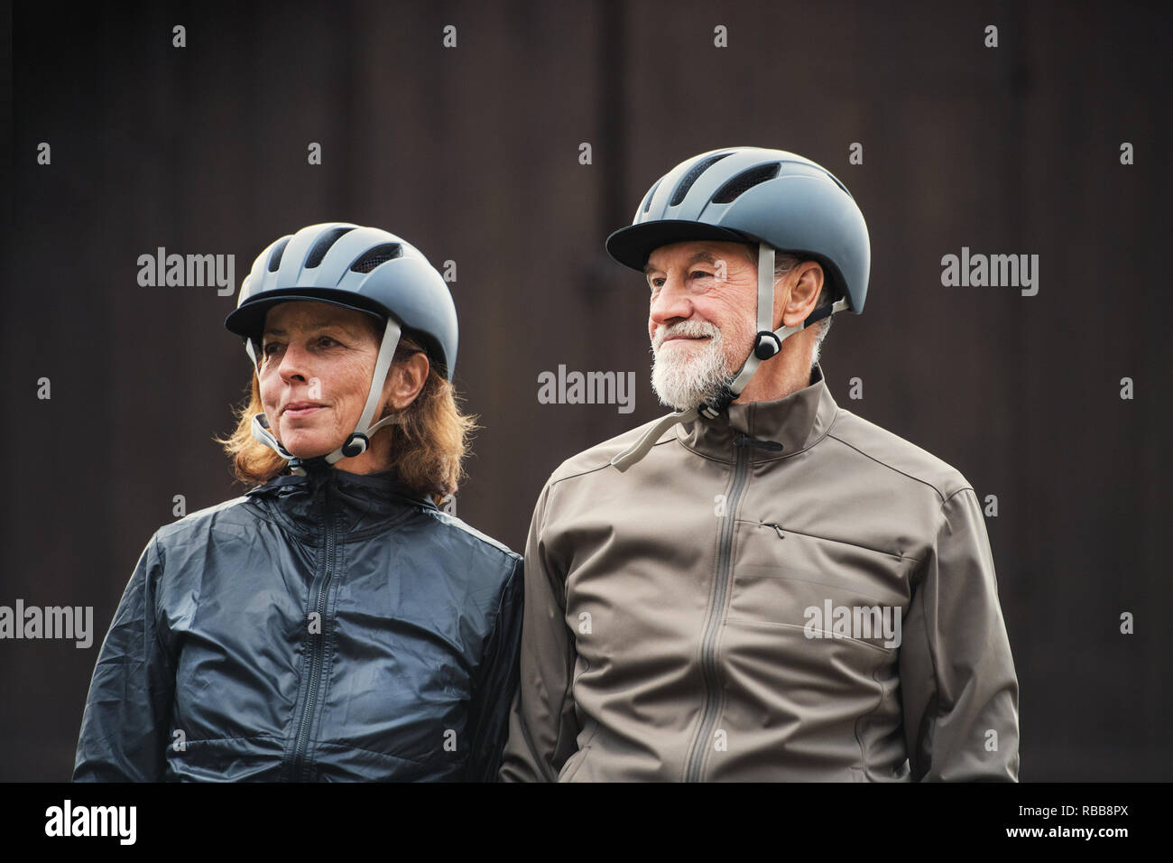 Senior couple avec casque de vélo debout à l'extérieur contre un arrière-plan sombre. Banque D'Images