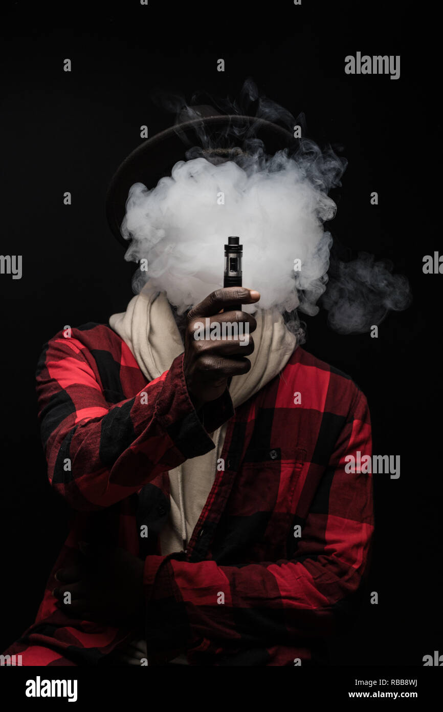 Jeune homme africain avec nuage de fumée sur le visage vape cigarette  électronique à l'aide d'un nuage de fumée, de trucs et astuces mod vaping  device arrière-plan Photo Stock