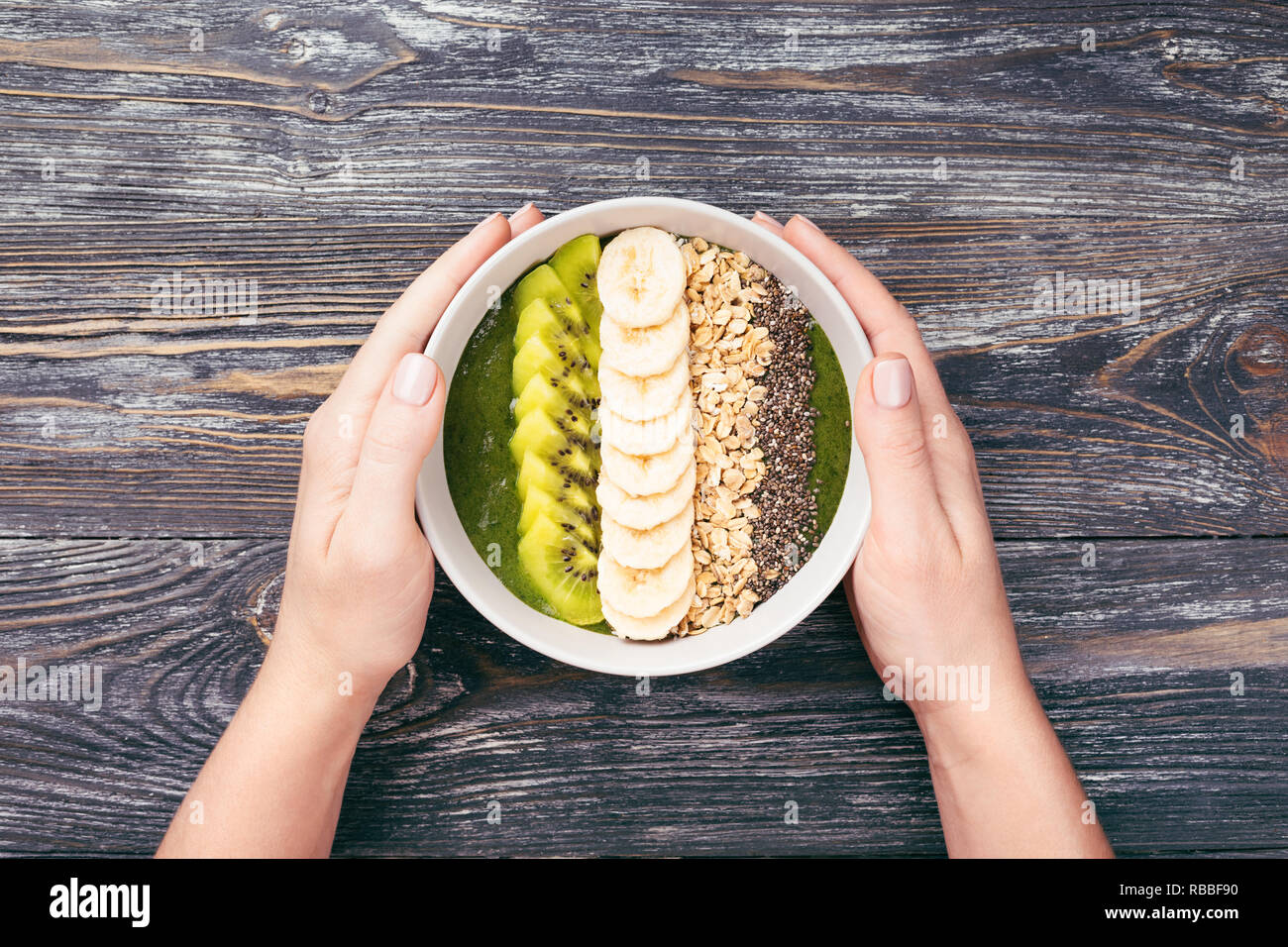 Woman's hand holding green smoothie épinards bol avec des céréales, graines de citrouilles, les bananes et les graines de quinoa sur table en bois. L'alimentation saine, vue du dessus, style rustique. Banque D'Images