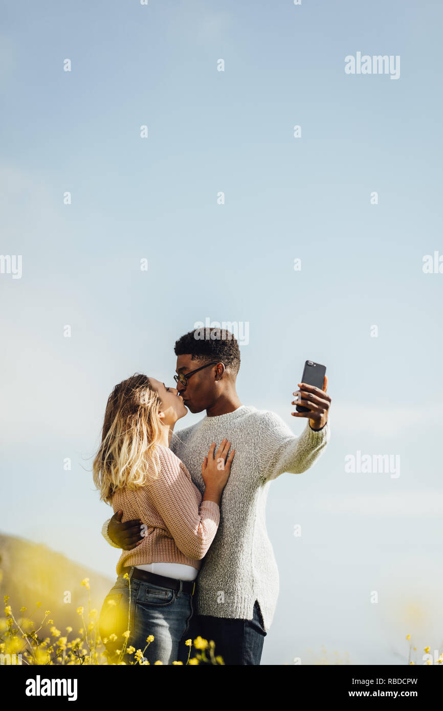 Couple interracial aimant et taking self portrait en extérieur. Couple romantique avec selfies leur téléphone mobile. Banque D'Images