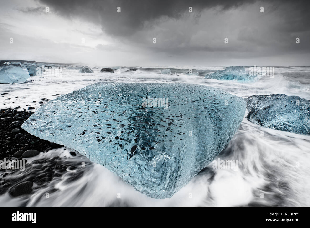 Gros bloc de glace dans les tons de bleu sur une plage avec de fortes vagues, le mouvement de l'eau peut être vu (longue exposition, traces de mouvement), au-dessus d'un contraste Banque D'Images